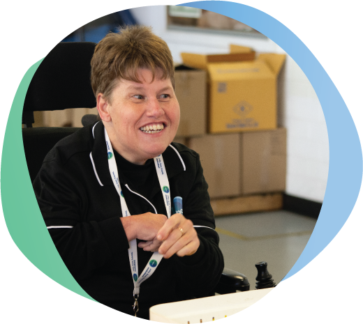 Person with disability wearing mobo shirt, smiling, sitting at workstation in powered wheelchair.