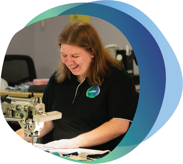 Person with disability wearing mobo shirt, smiling while working on sewing machine.