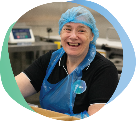 Person with disability wearing mobo shirt, apron and hairnet working on weighing and packaging goods in sterile food room.