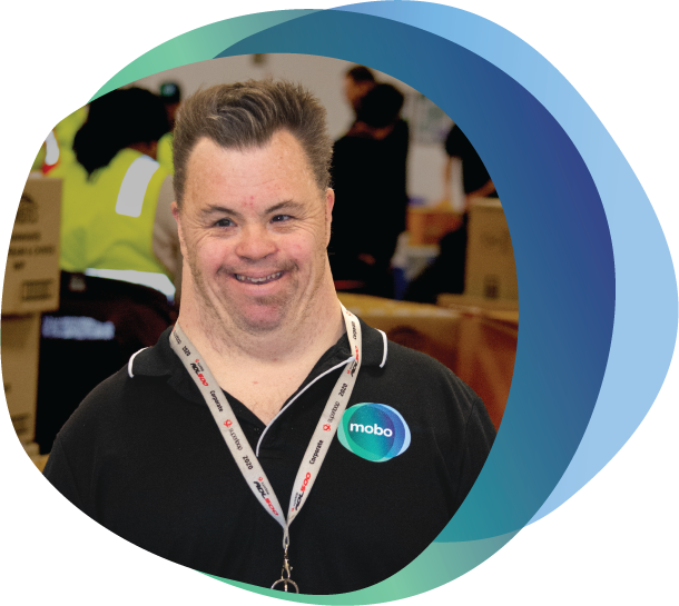Person with a disability wearing mobo shirt smiling at camera, with packaging work in the background.