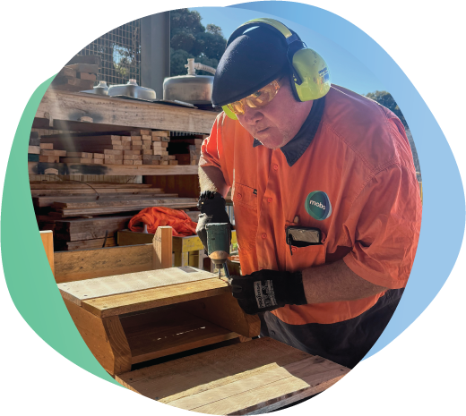 Person in orange Hi Vis shirt, safety glasses and ear protection using a drill, constructing a wooden box.